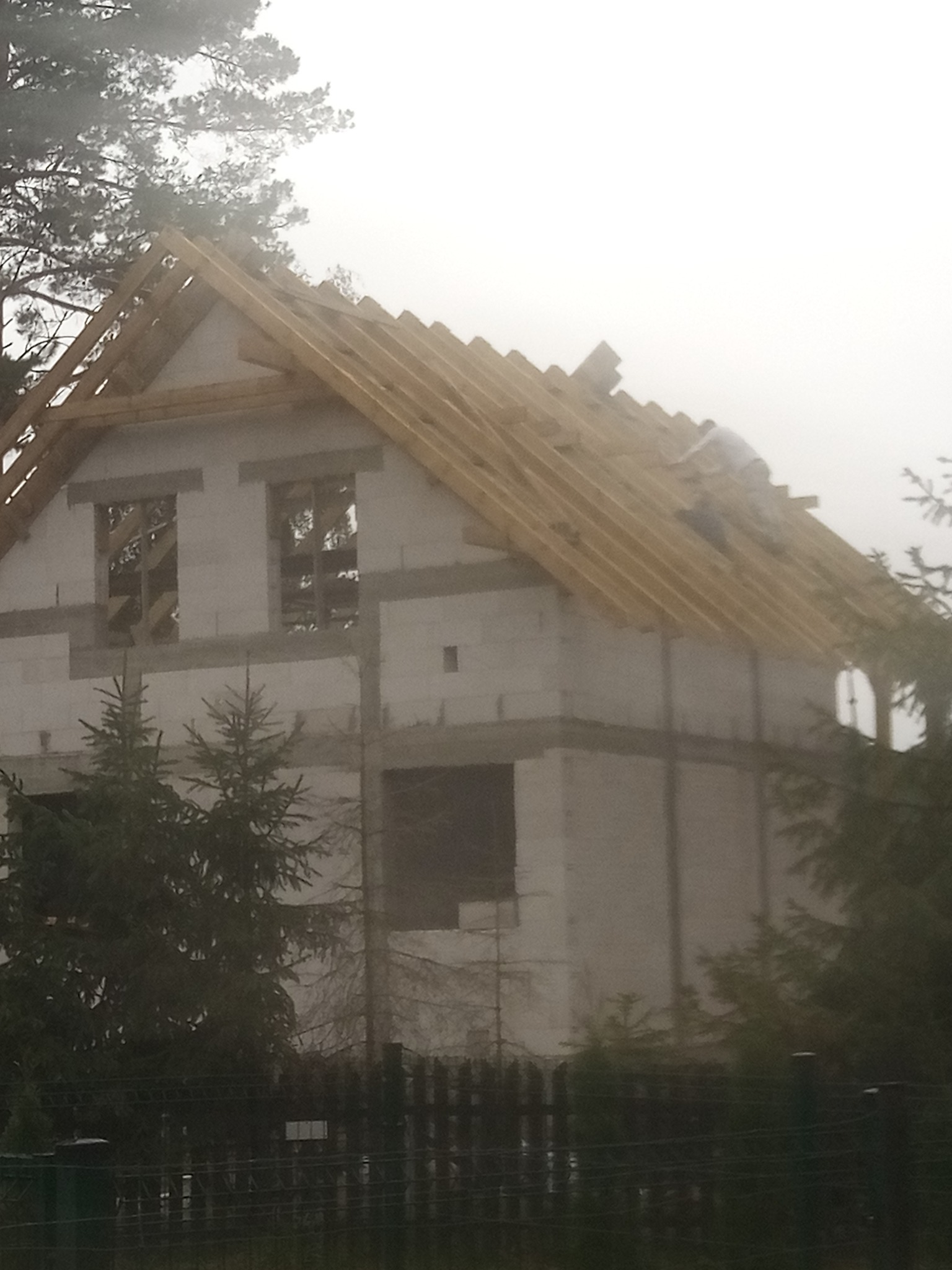 Dom w trakcie budowy w mglisty dzień. Widoczna konstrukcja więźby dachowej i ściany z pustaków. Na dachu pracuje osoba. Ujęcie zza ogrodzenia.