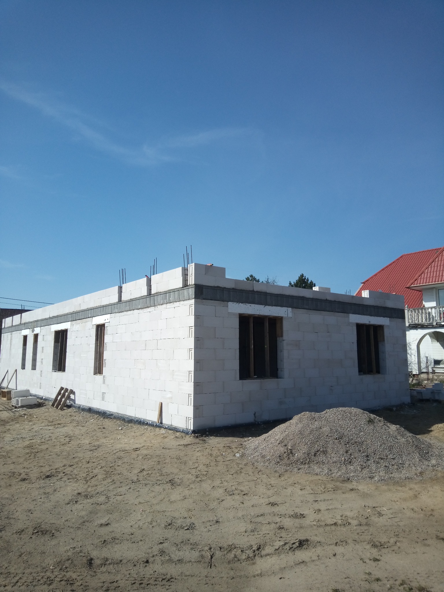 Niski budynek w trakcie budowy z białych bloczków, widoczne otwory okienne i wystające pręty zbrojeniowe. Na pierwszym planie hałda żwiru, w tle dom z czerwoną dachówką.