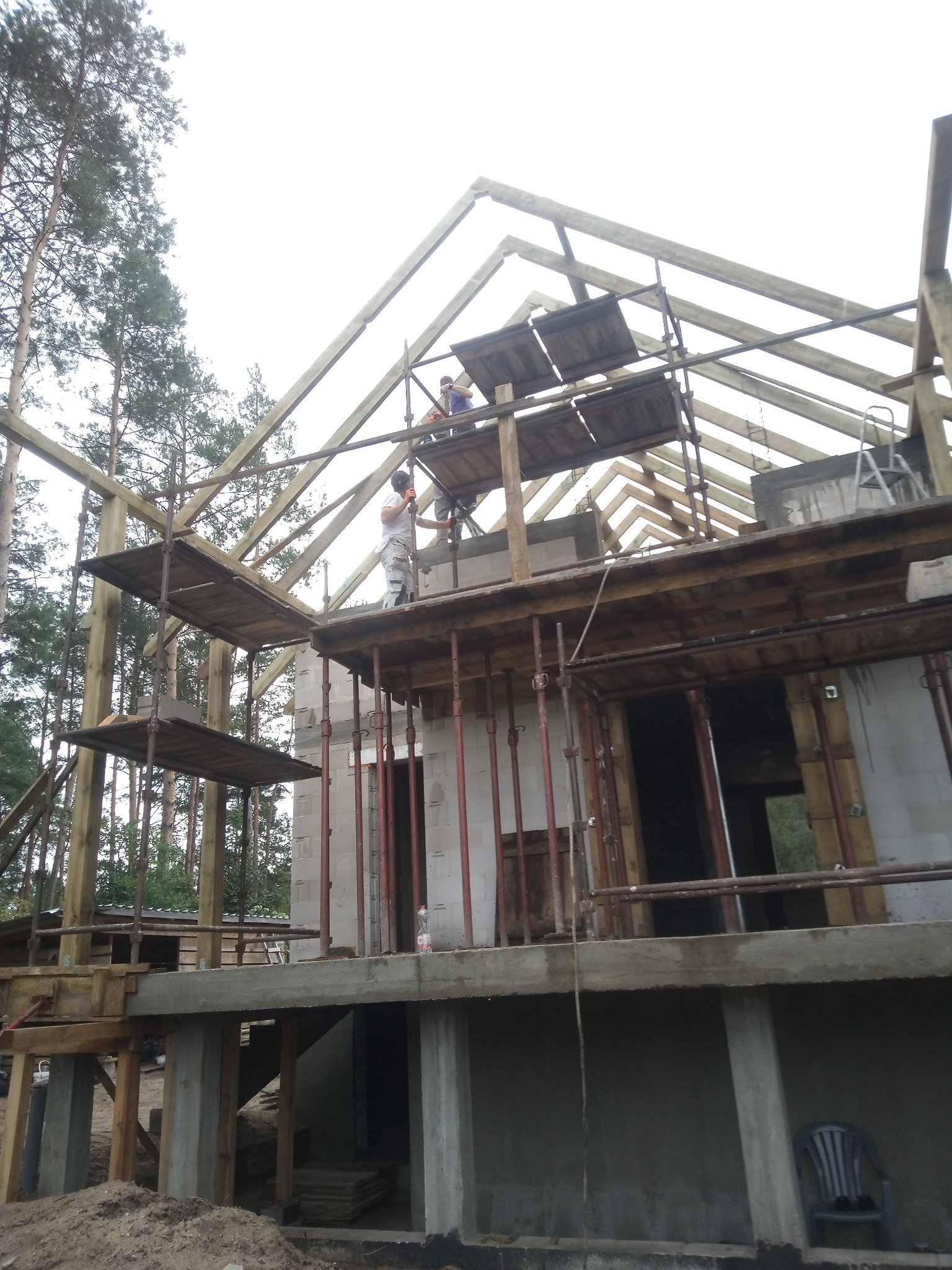 Budowa domu w trakcie: widoczna konstrukcja więźby dachowej z drewnianych belek, rusztowania i pracujący robotnicy. Ściany z betonu komórkowego.