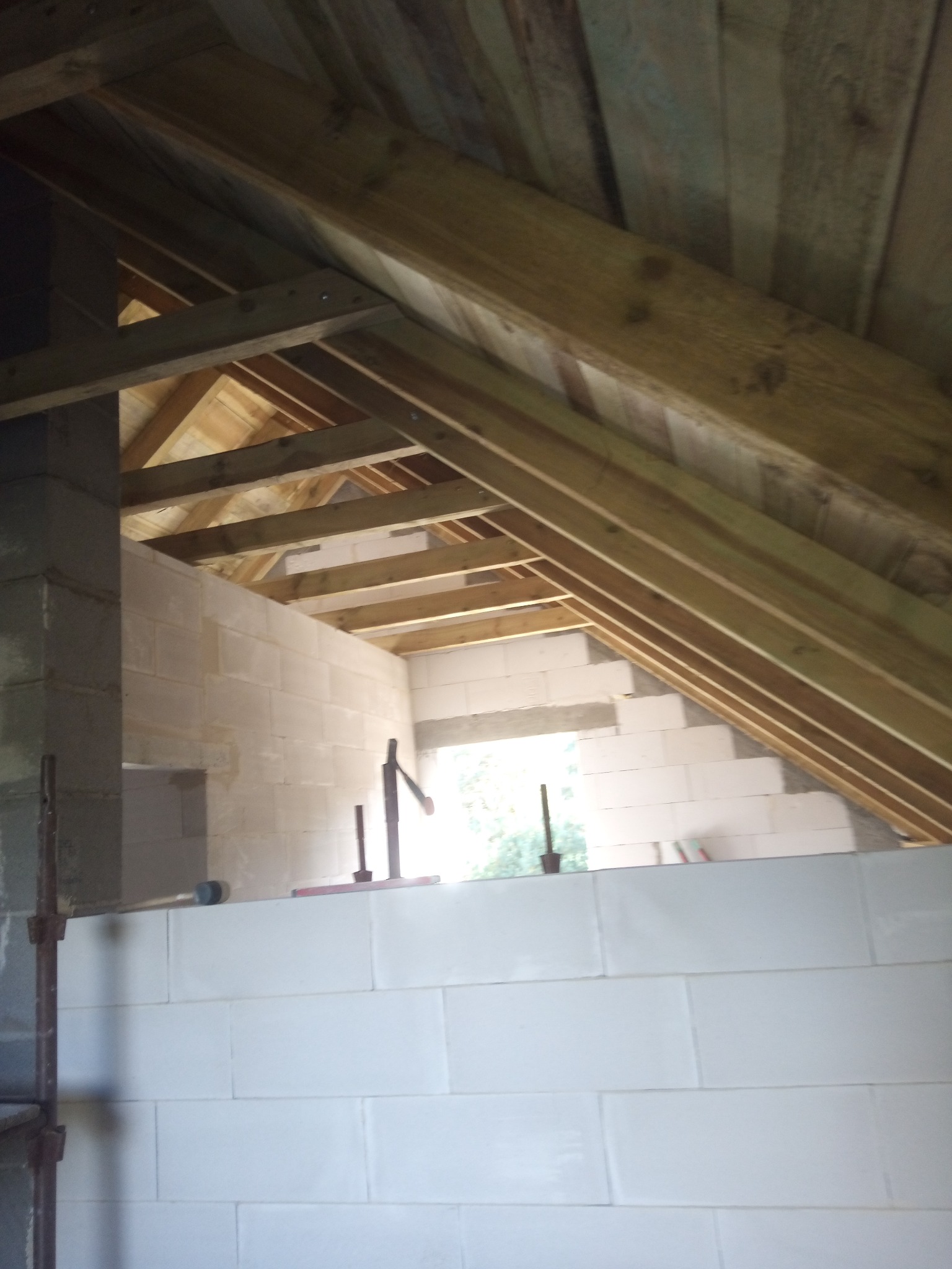 Wnętrze domu w trakcie budowy: białe ściany z betonu komórkowego, drewniana więźba dachowa, widoczne narzędzia murarskie. Surowy stan, jasne światło wpadające przez otwory okienne.