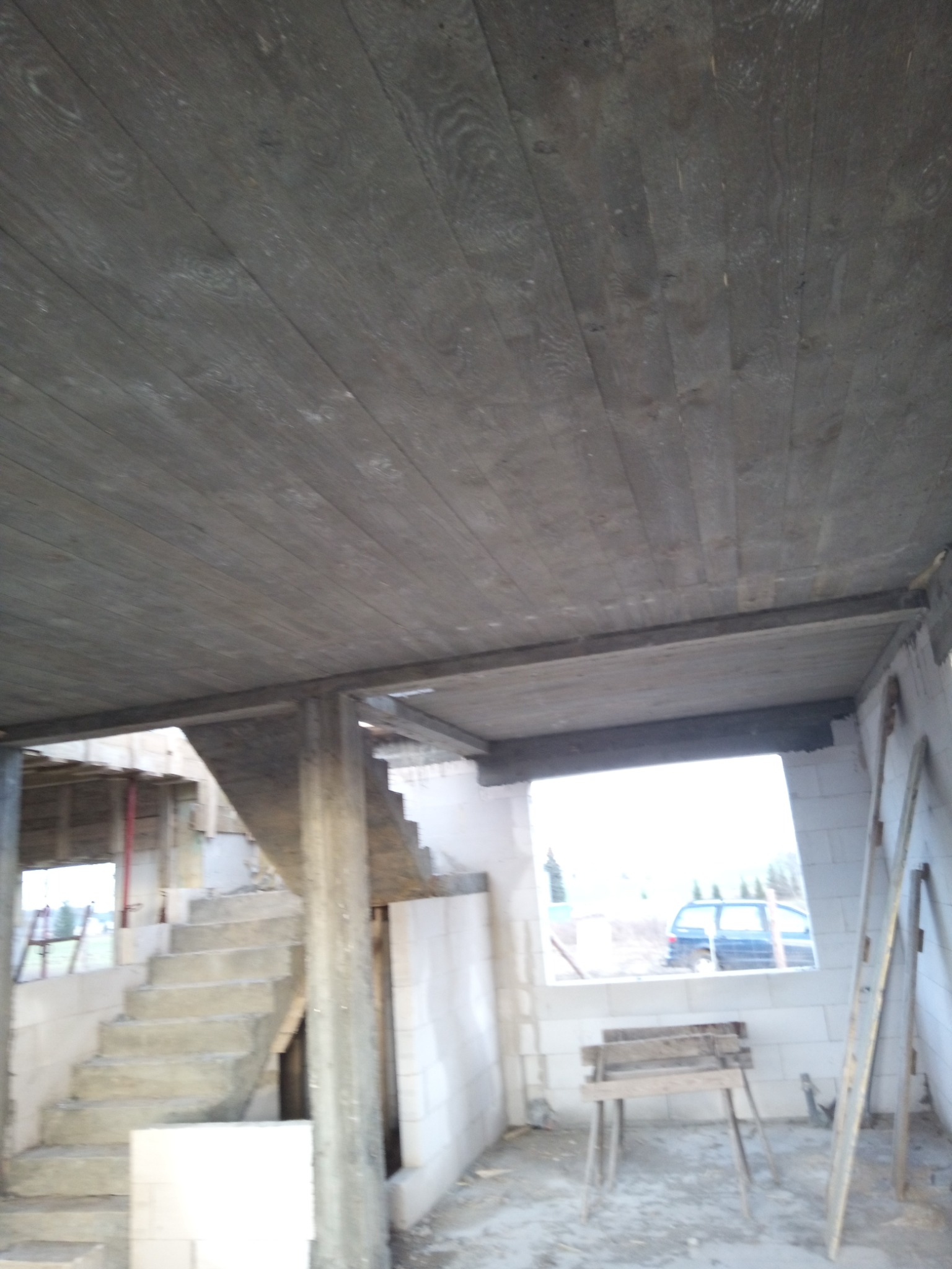 Surowy strop z desek w nowym domu, widoczne betonowe schody i pustaki. W tle okno z widokiem na zewnątrz i zaparkowany samochód. Wnętrze w stanie deweloperskim.