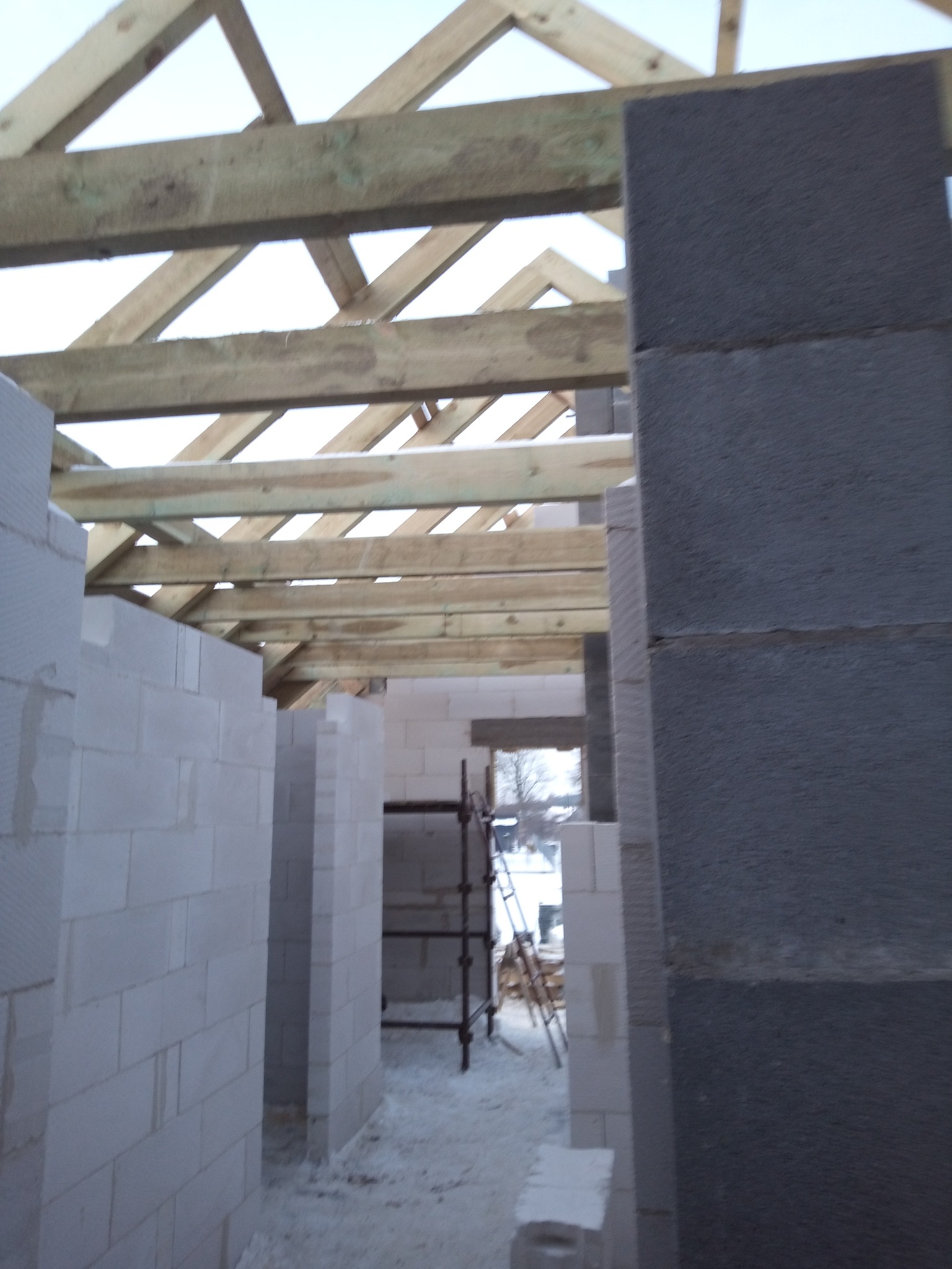 Wnętrze surowego domu w budowie: ściany z białych i szarych bloczków, drewniana więźba dachowa, widoczne rusztowania w głębi. Stan surowy otwarty.