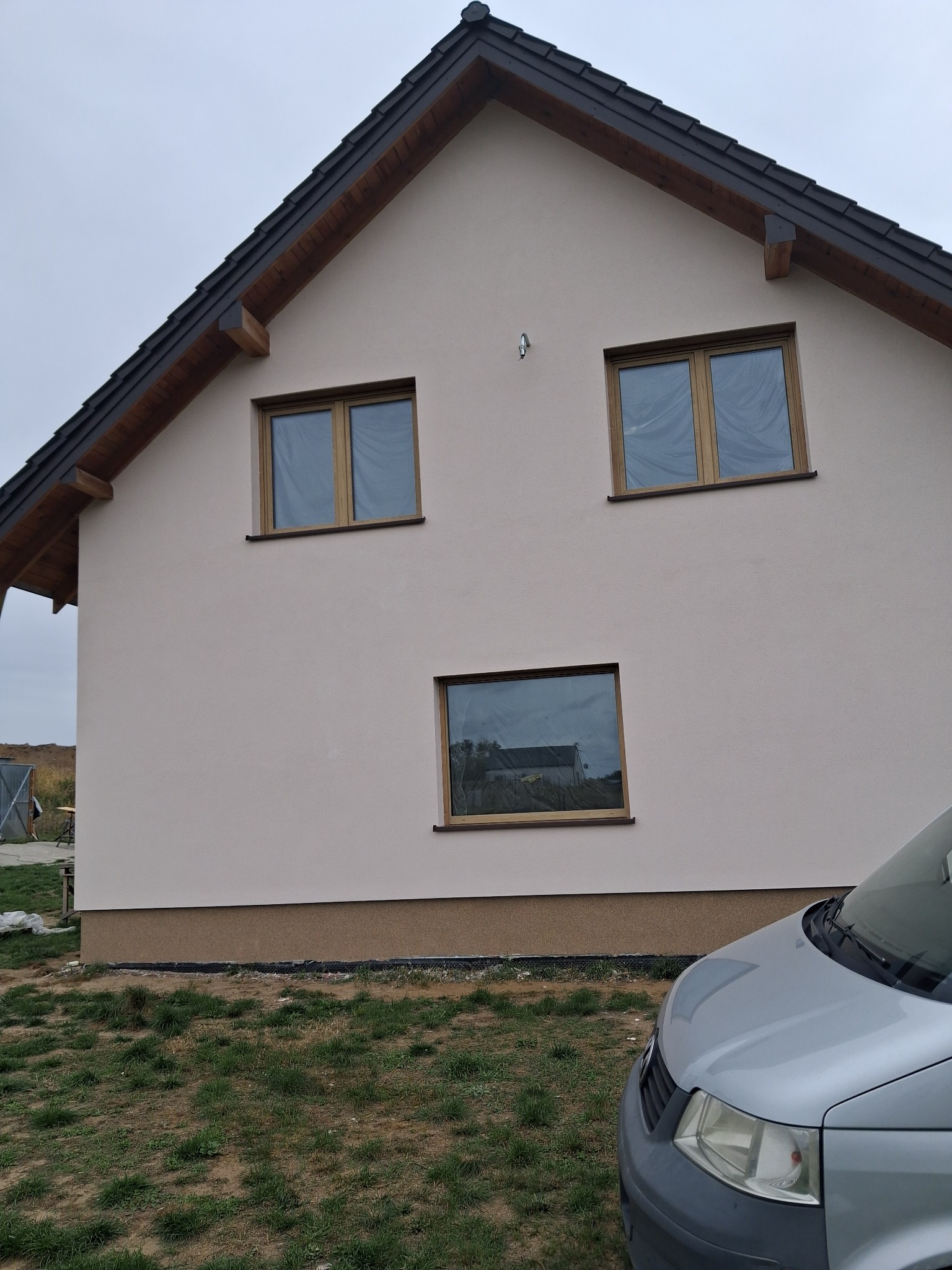 Jasny dom z ciemną więźbą dachową i oknami zabezpieczonymi folią, widziany z boku, z fragmentem srebrnego samochodu na pierwszym planie i pochmurnym niebem w tle.