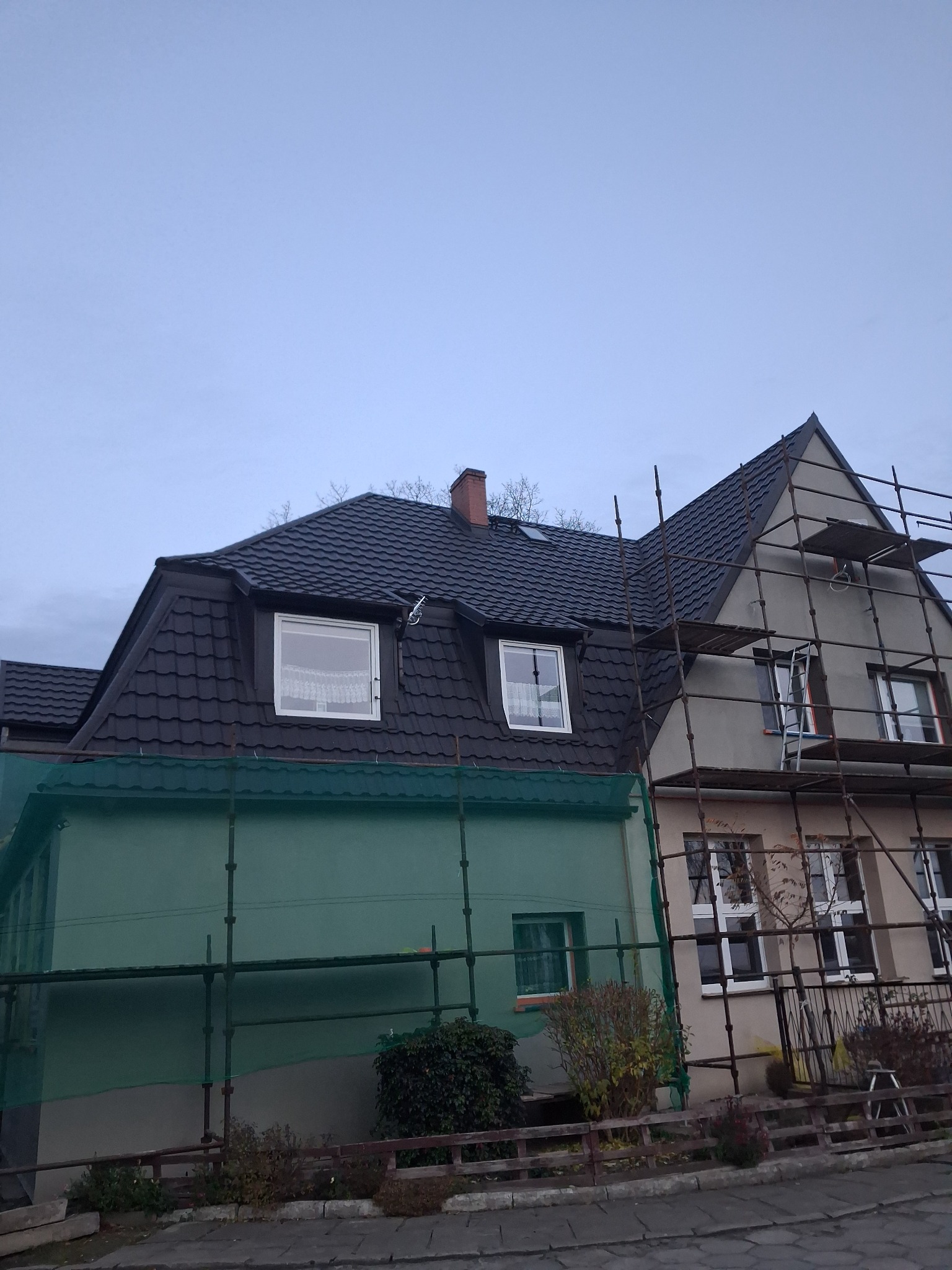 Dom w trakcie prac elewacyjnych: część budynku z nową, ciemną dachówką i oknami, druga część w rusztowaniach, z szarą elewacją. Fragment osłonięty zieloną siatką.