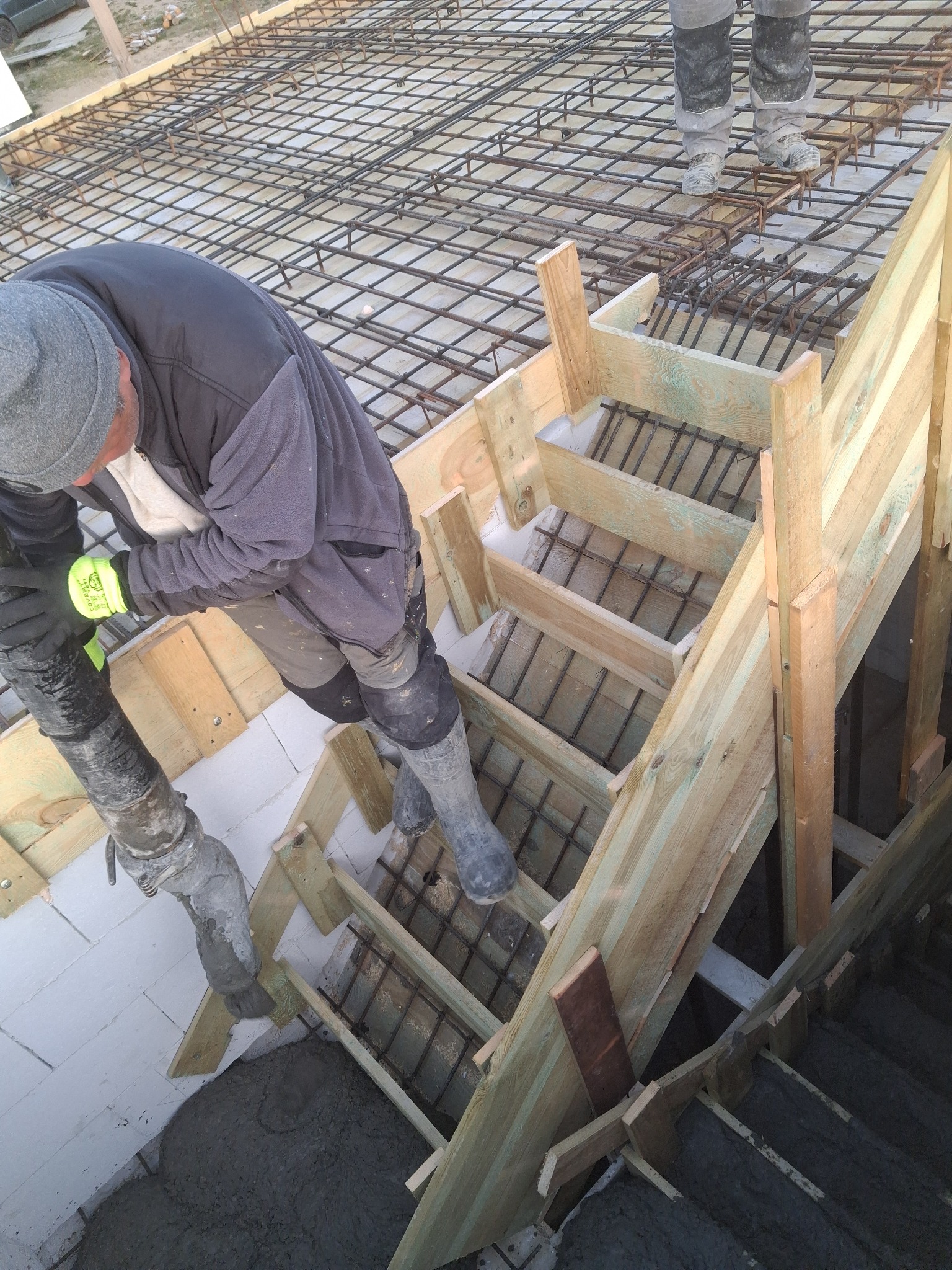 Zalewanie betonem schodów w szalunku zbrojonym stalą. Pracownik w kombinezonie i rękawicach obsługuje pompę do betonu. Widoczne elementy konstrukcji i zbrojenia.