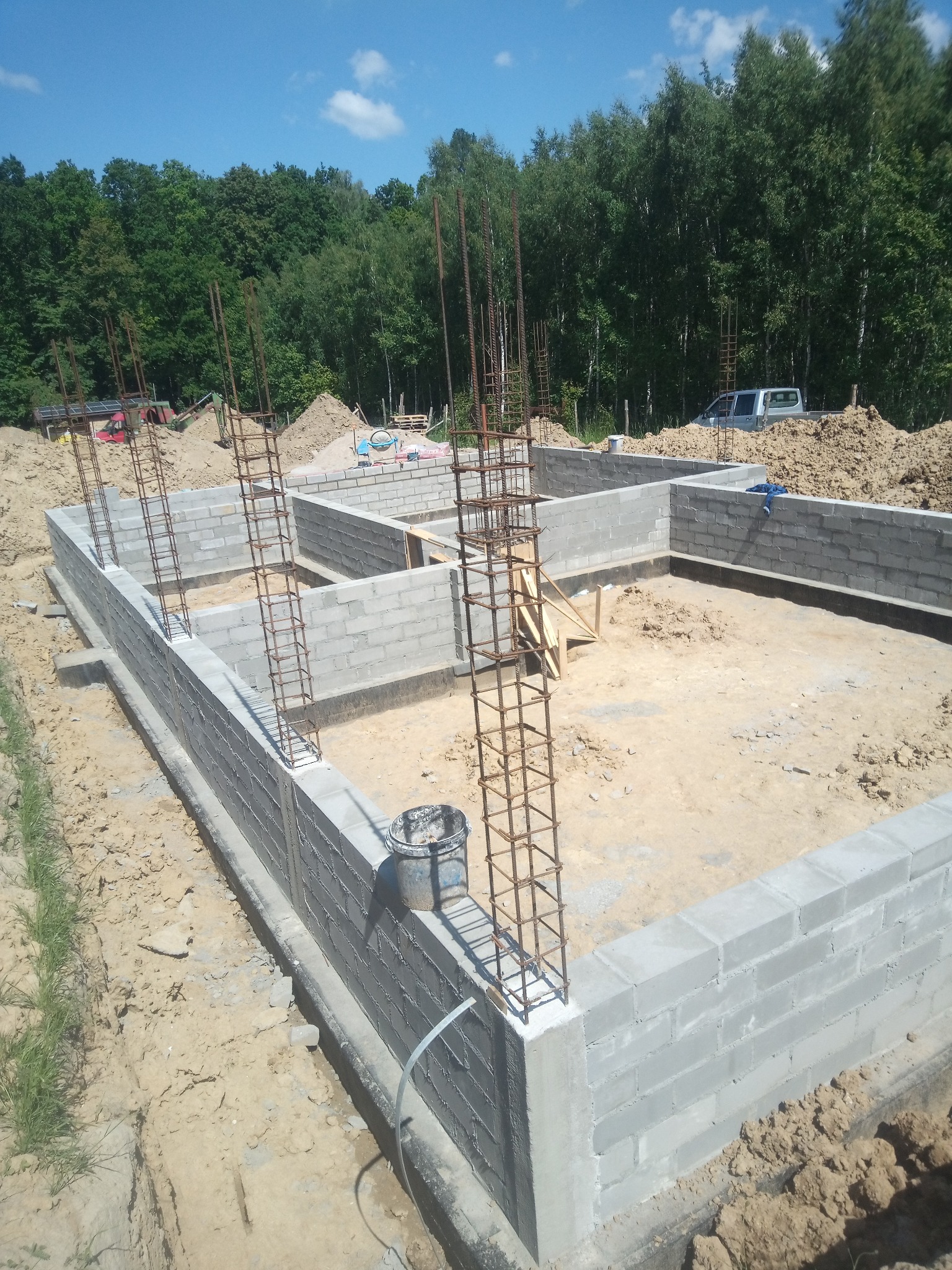 Fundamenty z bloczków betonowych i zbrojenia pionowego na tle zieleni. Widoczny etap budowy z wyraźną strukturą bloczków i detalami zbrojenia.