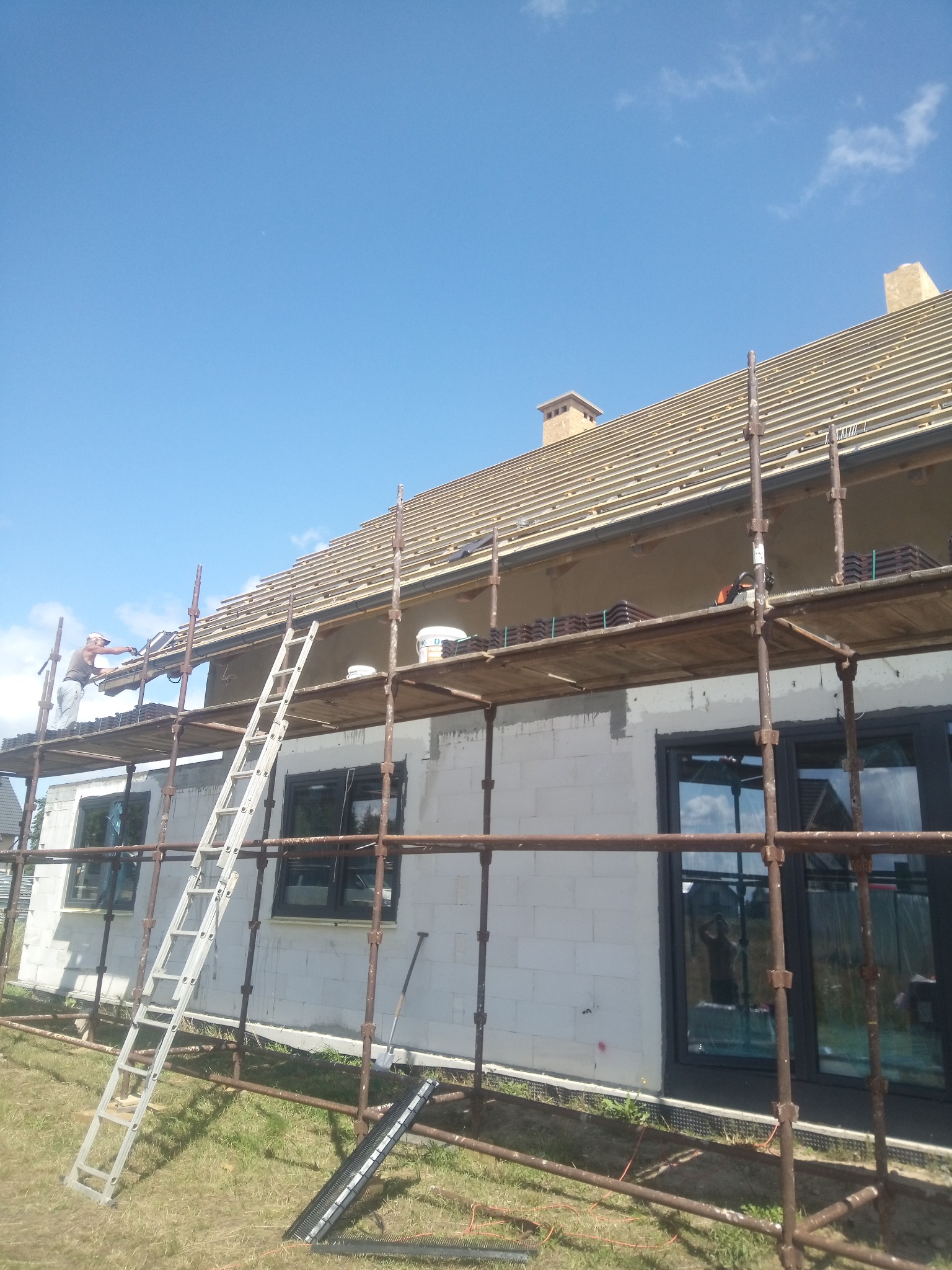 Budowa domu z białego bloczka: murarz na rusztowaniu układa dachówkę, widoczna drabina i okna z ciemnymi ramami. Dach w trakcie krycia, błękitne niebo w tle.