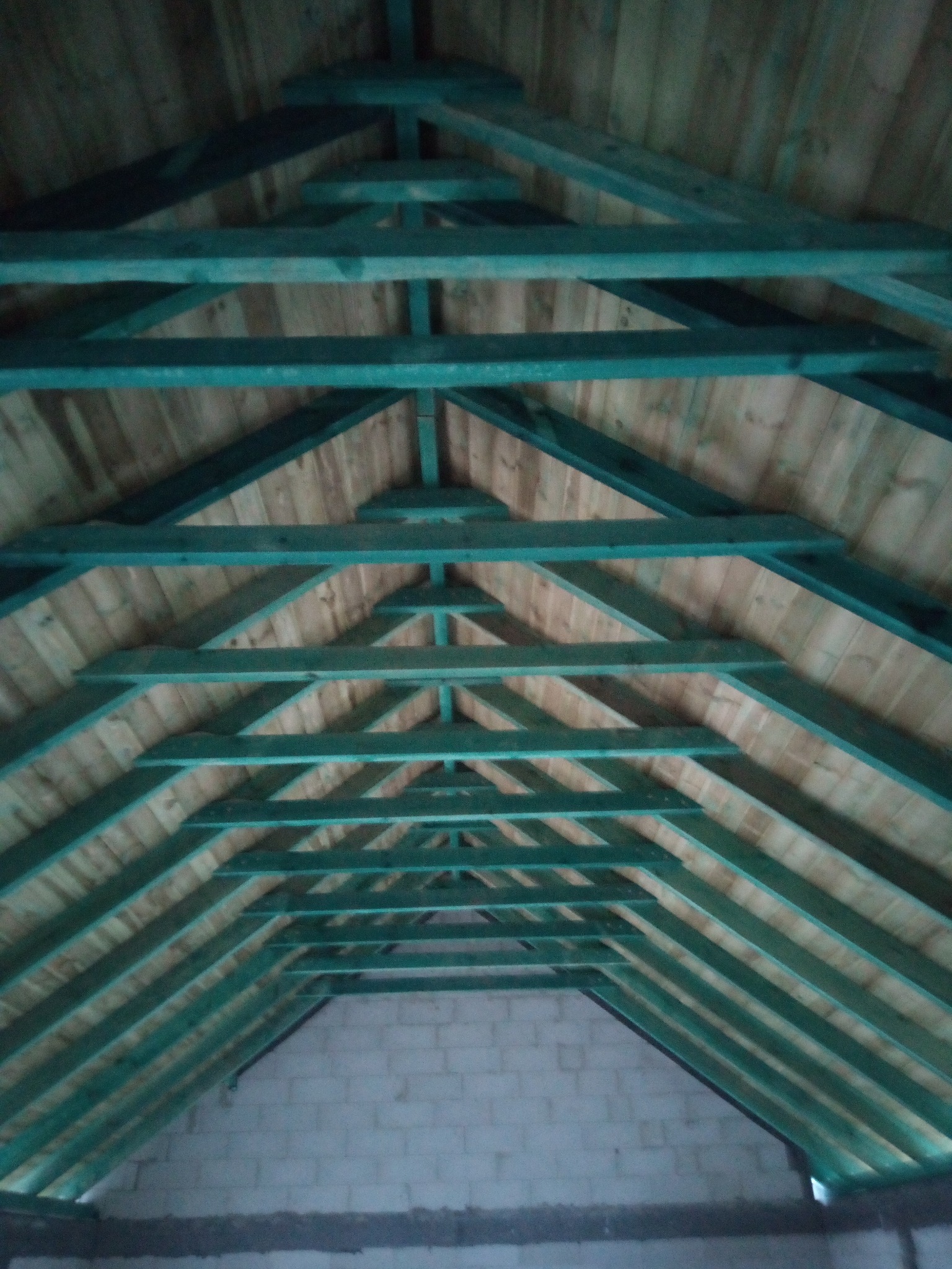 Drewniana więźba dachowa z zielonymi elementami konstrukcyjnymi, widok od dołu, z fragmentem murowanej ściany w tle. Układ belek tworzy symetryczny wzór trójkątów.