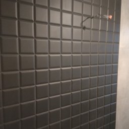 Kazimierz Duch remonty i wykończenia łazienek - Ściana wyłożona ciemnymi, kwadratowymi płytkami 3D z widocznym przewodem wystającym ze ściany.