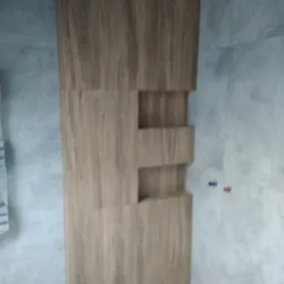 Wykończona ściana w łazience z drewnopodobną zabudową z wnękami, na tle szarych płytek i instalacji wodnej z niebieską i czerwoną zaślepką.