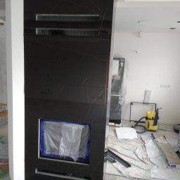 Kazimierz Duch remonty i wykończenia łazienek - Ściana wykończona czarnymi płytami z imitacją marmuru, z wnęką zabezpieczoną folią, widoczny odkurzacz na podłodze w trakcie prac wykończeniowych.