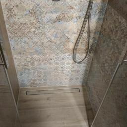 Kazimierz Duch remonty i wykończenia łazienek - Kabina prysznicowa z odpływem liniowym, wykończona płytkami w odcieniach beżu i szarości z dekoracyjnym wzorem na ścianach i imitacją drewna na podłodze.