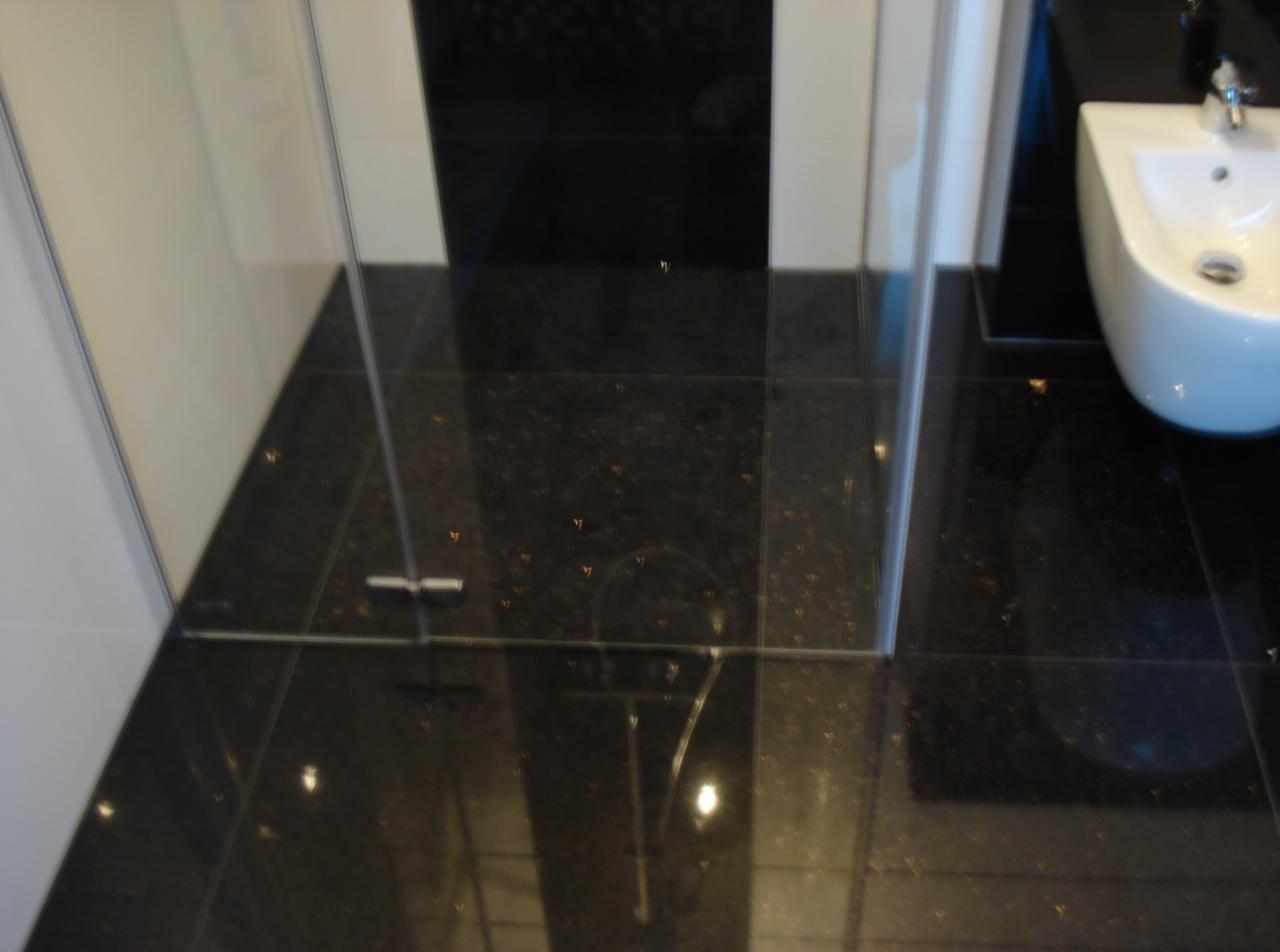 Nowoczesna łazienka z czarną, błyszczącą posadzką i białym bidetem. Szklana kabina prysznicowa z widocznym odpływem liniowym.