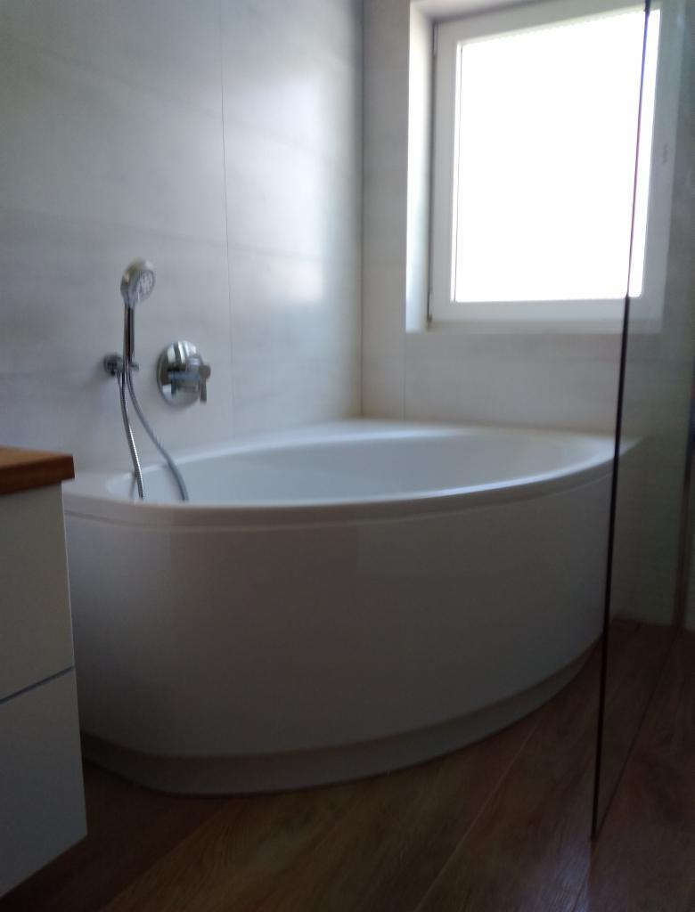 Biała, owalna wanna z zamontowaną baterią i słuchawką prysznicową na tle jasnych, marmurkowych płytek w łazience z drewnopodobną podłogą i oknem.