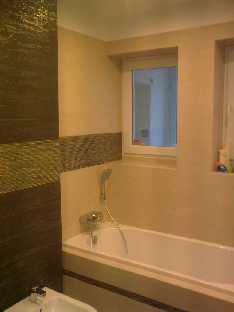 Beżowo-brązowa łazienka z wanną i bidetem. Na ścianach dekoracyjne płytki i mozaika. Widoczne okno z białym obramowaniem, armatura łazienkowa.