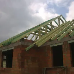 Niedokończony budynek z czerwonej cegły z drewnianą konstrukcją dachu na tle pochmurnego nieba. Widoczne okna bez szyb.