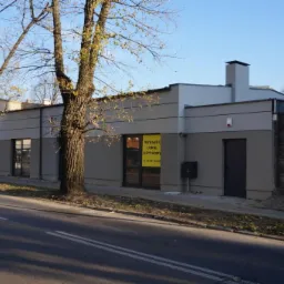 Parterowy budynek w odcieniach szarości z płaskim dachem i kominem, widoczny z ulicy. Na oknach ogłoszenie 'Wynajmę lokal użytkowy'.