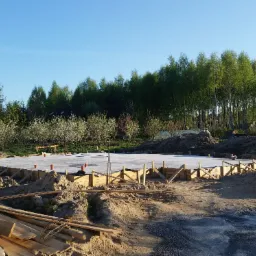 Wylana płyta fundamentowa z drewnianym szalunkiem na placu budowy w słoneczny dzień, w tle drzewa i zabudowania.