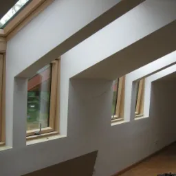 Poddasze z oknami dachowymi w drewnianych ramach, białe ściany i skośny sufit z belkami. Widok na zewnątrz przez szyby okien.