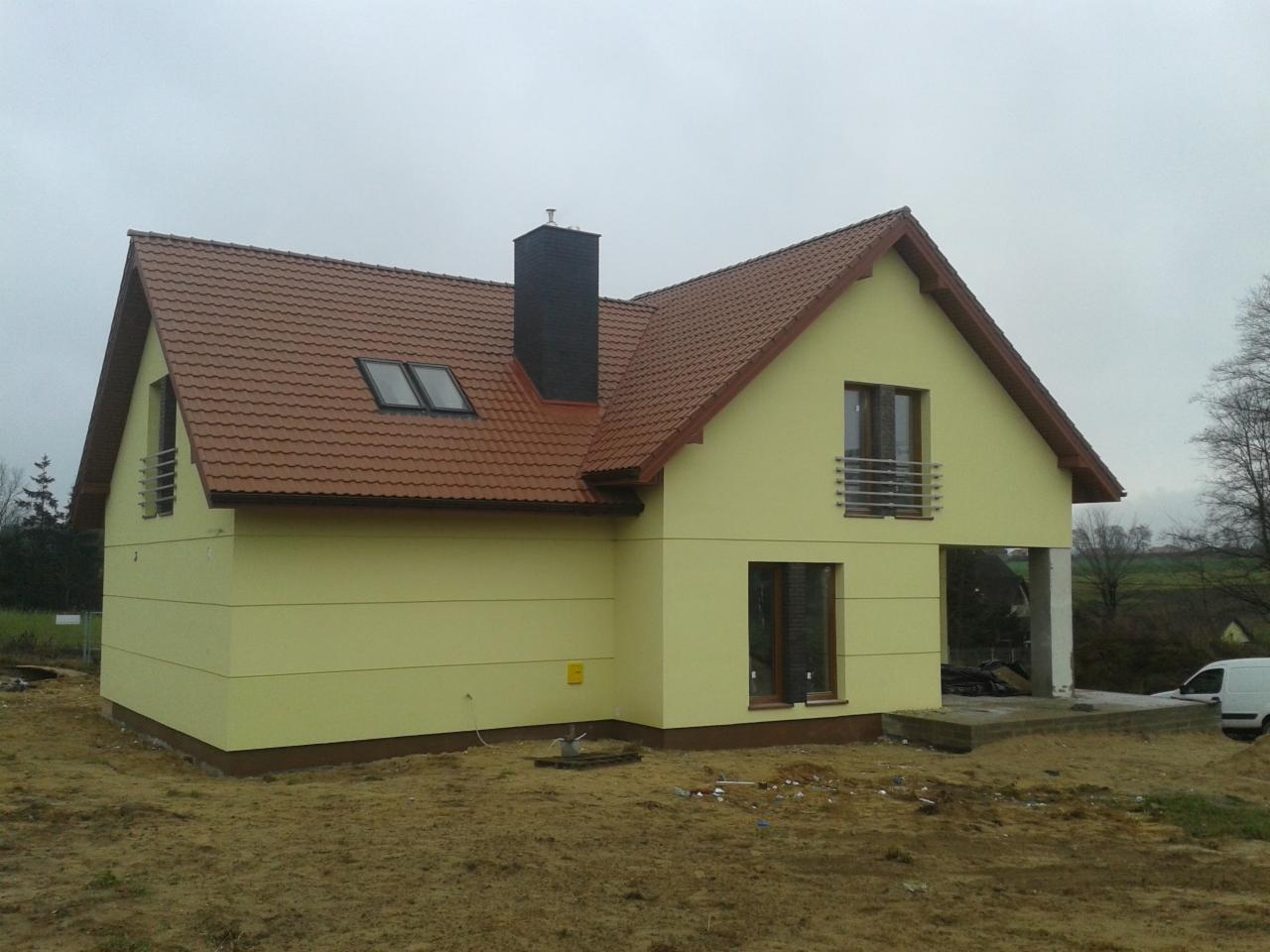 Żółty dom jednorodzinny z brązowym dachem i czarnym kominem. Widoczne okna dachowe i balkon z metalową barierką. Częściowo wykończony, w surowym otoczeniu.