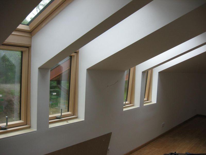 Poddasze z oknami dachowymi w drewnianych ramach, białe ściany i skośny sufit z belkami. Widok na zewnątrz przez szyby okien.