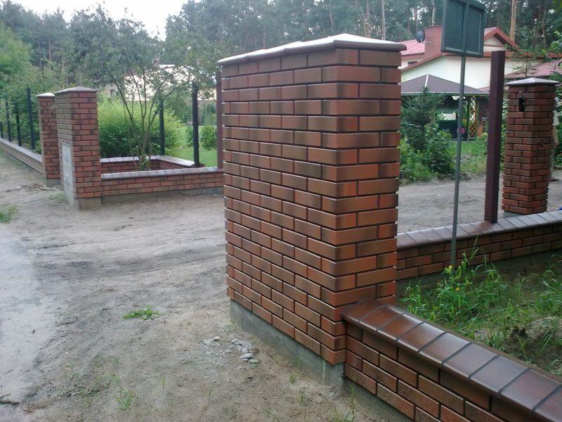 Nowoczesne ogrodzenie z czerwonej cegły klinkierowej z metalowymi przęsłami, widok z bliska na fragment ogrodzenia i słupki, teren posesji.