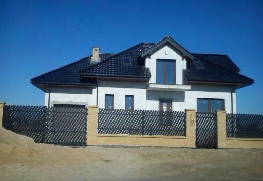 Dwukondygnacyjny dom z ciemnym dachem i drewnianym ogrodzeniem z numerem. Jasna elewacja, błękitne niebo w tle. Ujęcie z dołu.