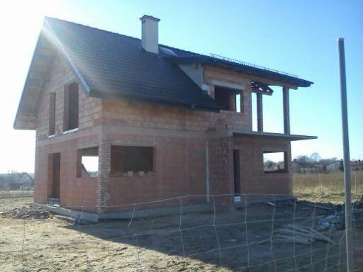 Dwukondygnacyjny dom w trakcie budowy z czerwonej cegły, z widocznym dachem i kominem na tle błękitnego nieba, ogrodzony siatką.