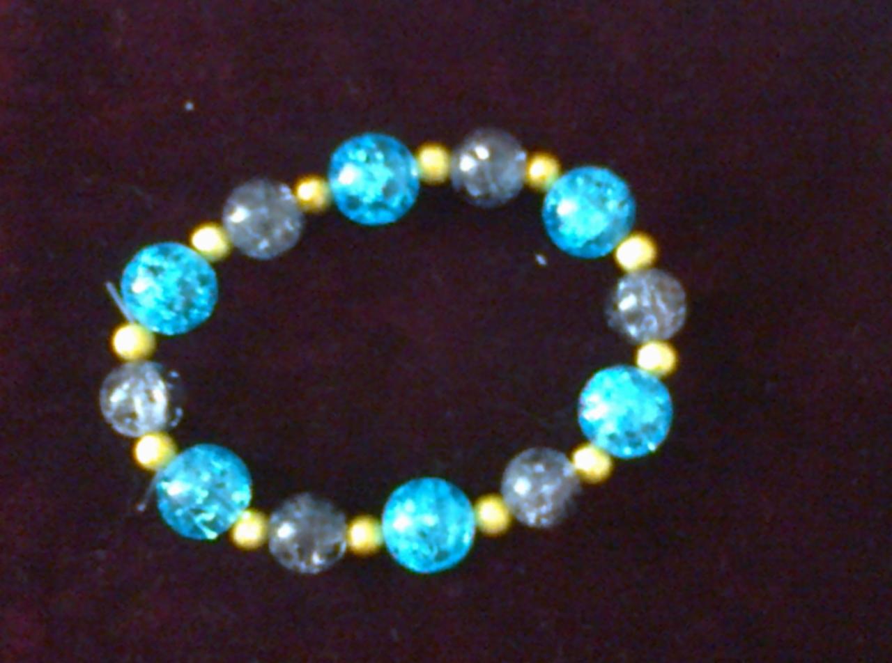 Elastyczna bransoletka z turkusowych i szarych koralików, oddzielonych małymi złotymi koralikami, leżąca na ciemnym, aksamitnym tle.