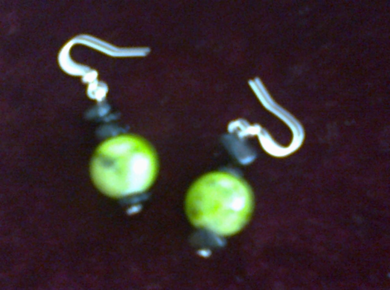 Dwa kolczyki wiszące z jasnymi, kulistymi, zielonymi koralikami i ciemnymi, nieregularnymi kamykami, zawieszone na srebrnych haczykach, na ciemnym, rozmytym tle.