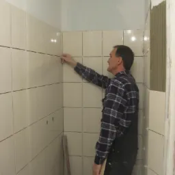 Układanie beżowych płytek w łazience w Grabno151. Fachowiec w kombinezonie dociska płytkę do ściany. Widoczne krzyżyki dystansowe i niezakończone fragmenty.