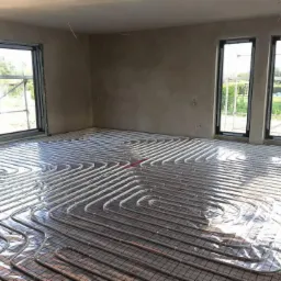 Instalacja ogrzewania podłogowego w surowym wnętrzu z dwoma oknami, widoczne rury grzewcze ułożone na siatce, ściany w stanie deweloperskim.