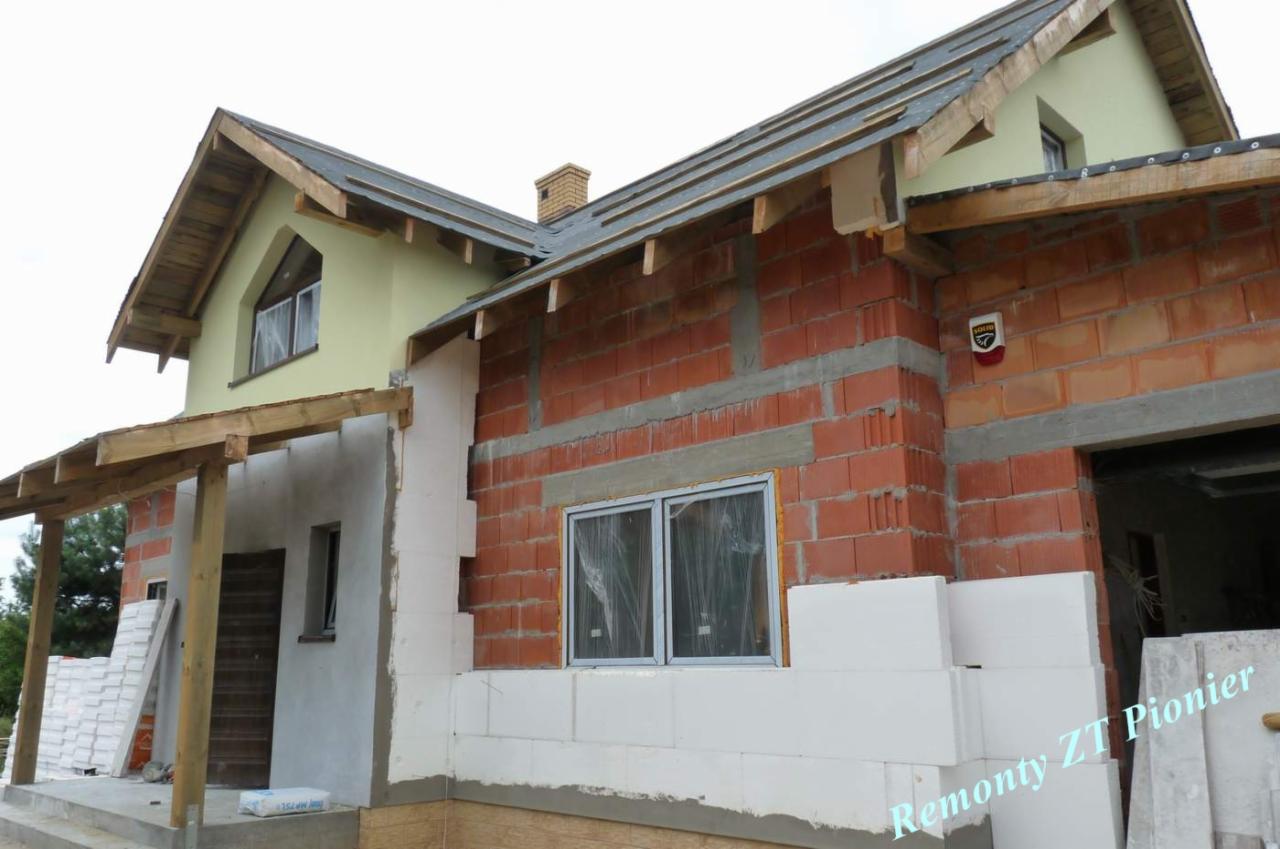 Budowa domu w trakcie: wznoszone ściany z czerwonej cegły i białego betonu komórkowego, widoczne okna z folią ochronną, drewniana konstrukcja zadaszenia.