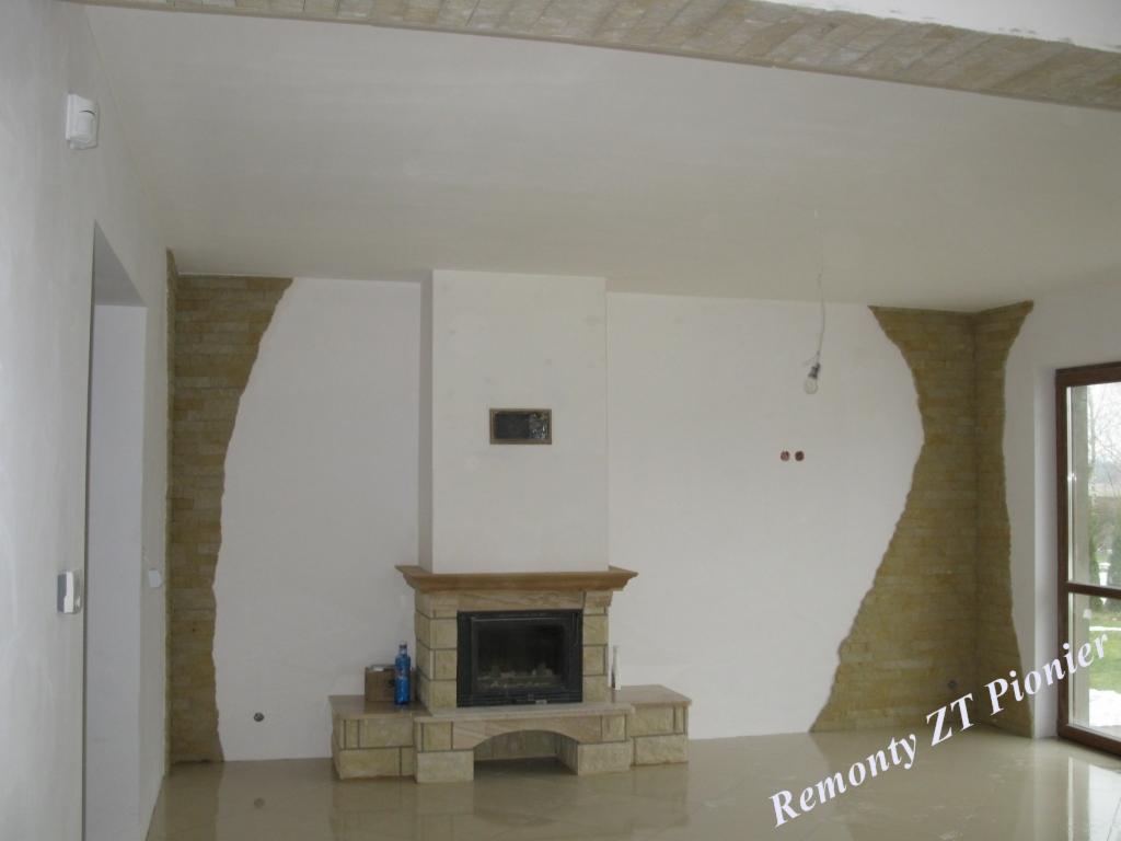 Salon po remoncie z kominkiem obłożonym kamieniem i fragmentami ścian z dekoracyjną cegłą. Widoczne ślady po pracach wykończeniowych na podłodze.
