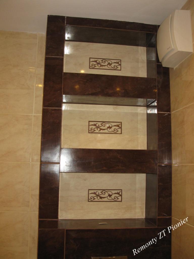 Narożna półka z płytek w łazience, z dekoracyjnymi wstawkami i wentylatorem na ścianie. Widoczne beżowe płytki na ścianach, brązowe obramowanie półki.