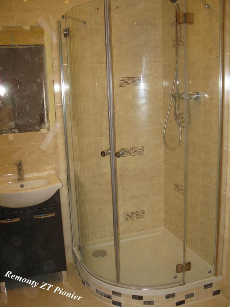 Wykończona łazienka z kabiną prysznicową, umywalką z szafką i beżowymi płytkami z dekorami. Widoczny fragment lustra. Ujęcie z niskiego kąta.