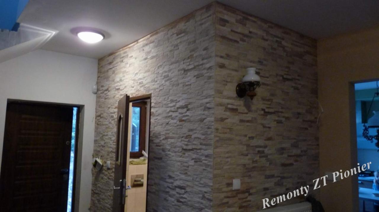 Wykończone wnętrze: ściana z dekoracyjnej cegły w odcieniach beżu i szarości, oświetlona kinkietem w stylu retro. Widoczne drzwi wejściowe i fragment sąsiedniego pomieszczenia.
