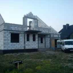 Wznoszenie murów domu z betonu komórkowego Ytong, widoczny częściowo ukończony szczyt i otwory okienne, na pierwszym planie taczka, w tle budynek mieszkalny i biały bus z otwartymi drzwiami.