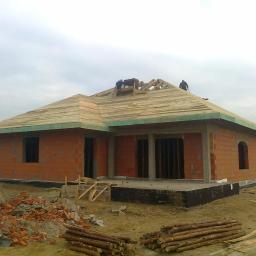 Zrb.budymex - Budowa domu jednorodzinnego z czerwonej cegły, widoczne deskowanie dachu, więźba dachowa w trakcie montażu i robotnicy na dachu, stosy drewna i gruzu na pierwszym planie.