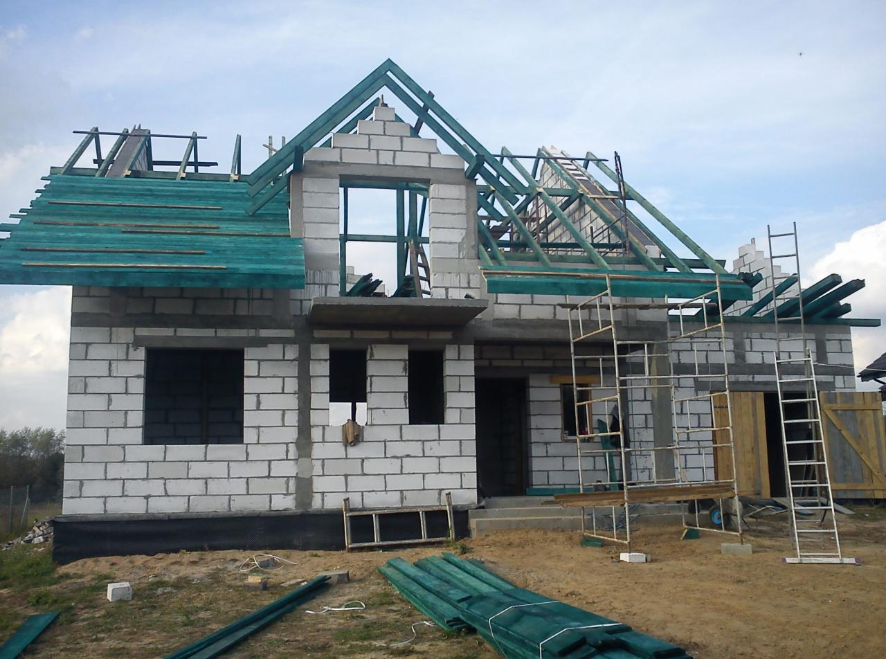 Wznoszenie ścian z bloczków betonu komórkowego i montaż konstrukcji więźby dachowej z zielonym deskowaniem na budowie domu jednorodzinnego.