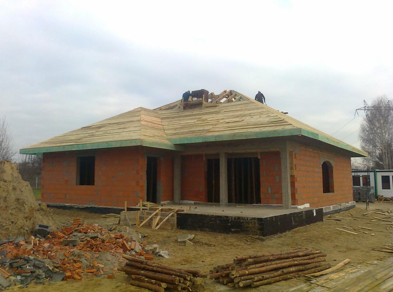 Budowa domu jednorodzinnego z czerwonej cegły, widoczne deskowanie dachu, więźba dachowa w trakcie montażu i robotnicy na dachu, stosy drewna i gruzu na pierwszym planie.