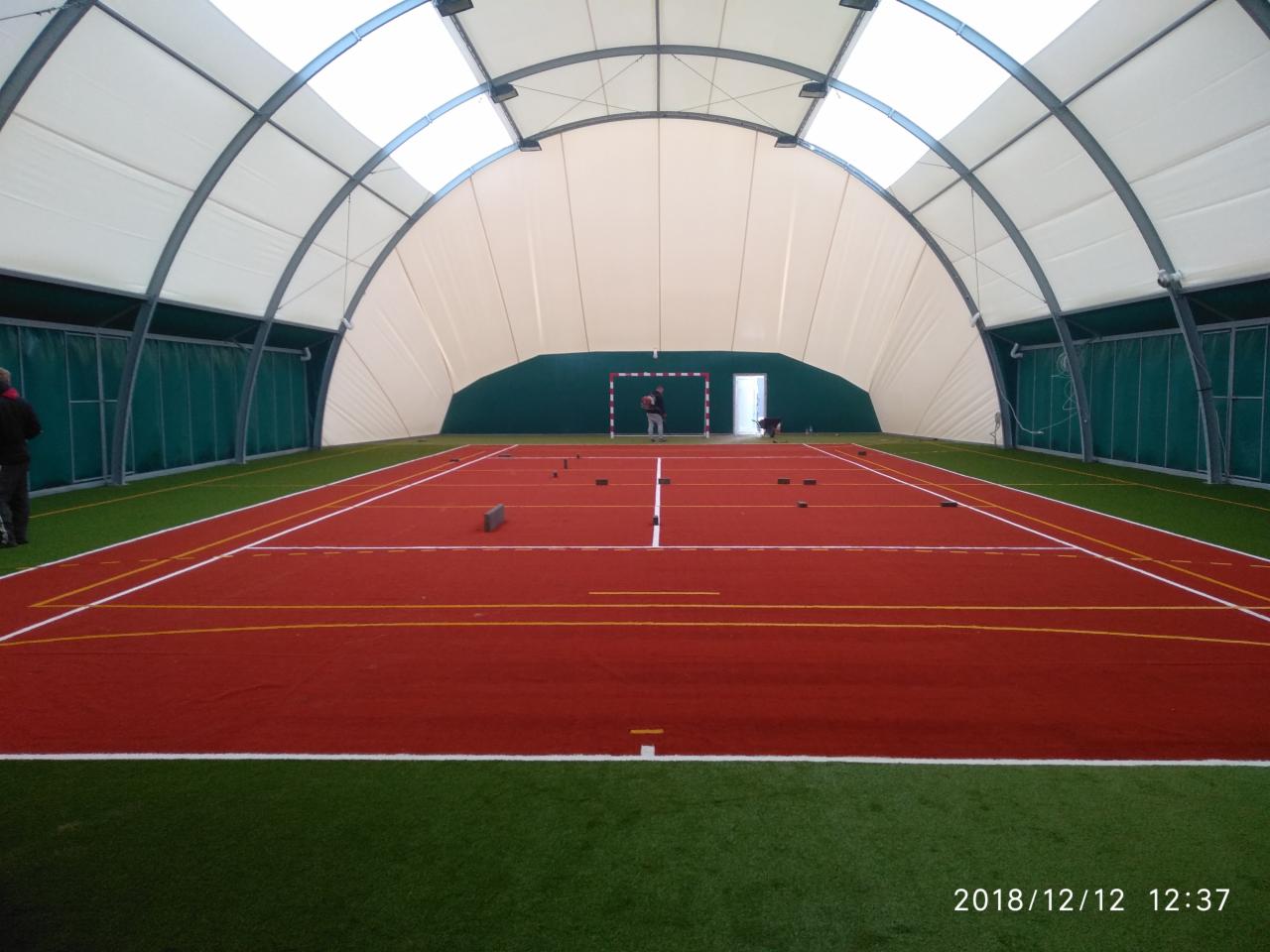 Wnętrze hali sportowej z czerwoną nawierzchnią boiska i zielonym obramowaniem z trawy syntetycznej, widoczne białe linie wyznaczające pole gry, w tle osoby i bramka.