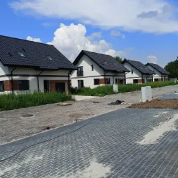 Szereg nowo wybudowanych, jednorodzinnych domów z czarnymi dachami i białymi elewacjami, widok z poziomu brukowanej drogi w trakcie układania, błękitne niebo z białymi obłokami w tle.