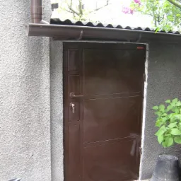 Brązowe, metalowe drzwi zewnętrzne z widocznym zamkiem i kluczem, zamontowane w szarej ścianie z fakturą, z rynną i fragmentem roślinności w tle.