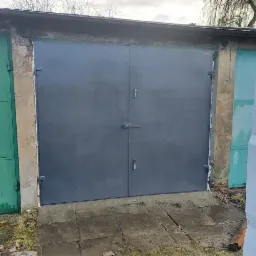 Szara, dwuskrzydłowa brama garażowa z zamkiem na klucz, świeżo osadzona w betonowej wylewce, widoczna na tle innych garaży i drzew bezlistnych.