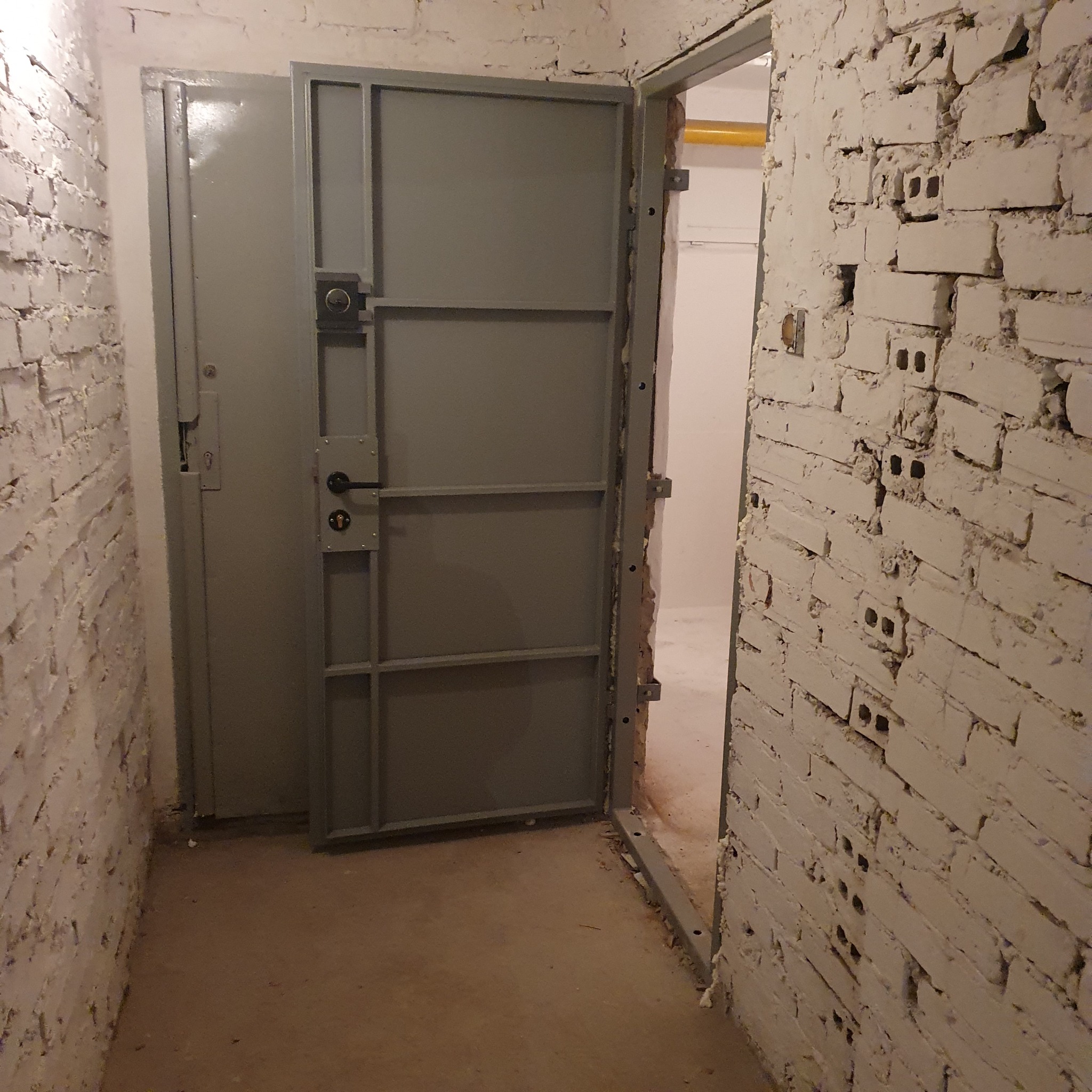 Otwarta, szara, metalowa brama z widocznym zamkiem i klamką, zamontowana w wejściu do pomieszczenia z pomalowanymi na biało ścianami z cegły dziurawki.