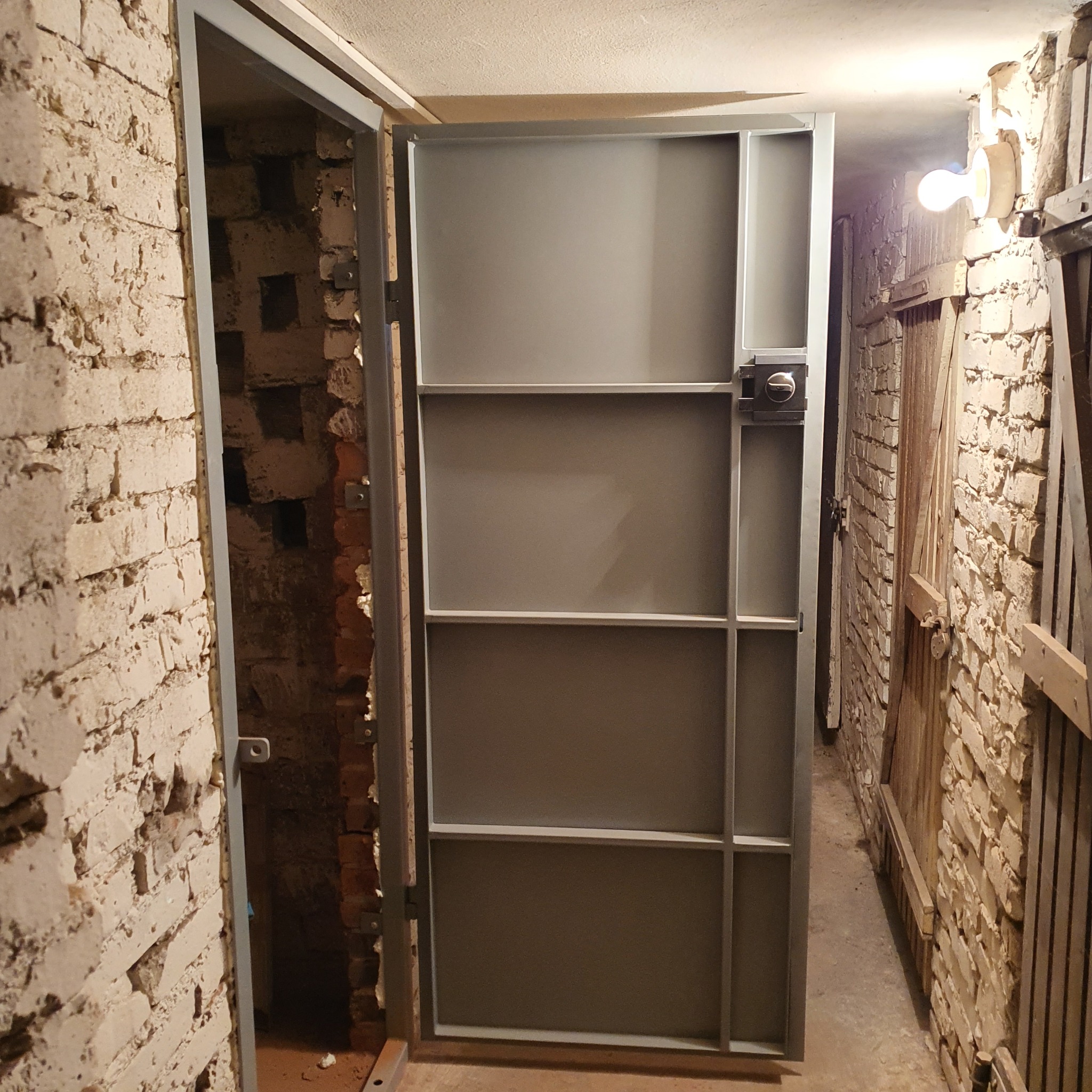Szara, metalowa krata drzwiowa z zamkiem, zamontowana w surowym, ceglanym pomieszczeniu piwnicznym z widoczną instalacją elektryczną.