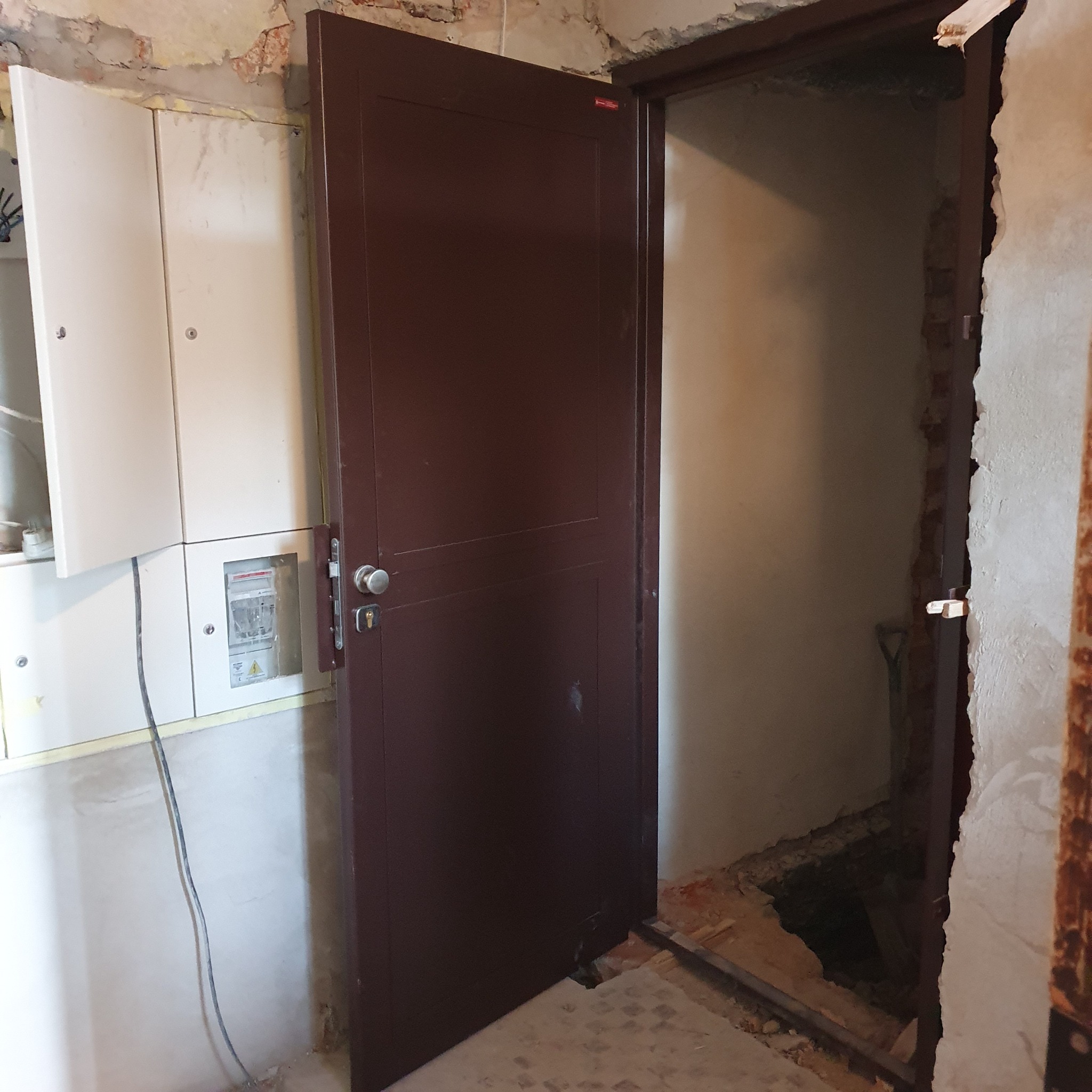 Brązowe drzwi z zamkiem i klamką, zamontowane w surowym otworze drzwiowym w trakcie remontu, widoczna skrzynka elektryczna na ścianie obok.