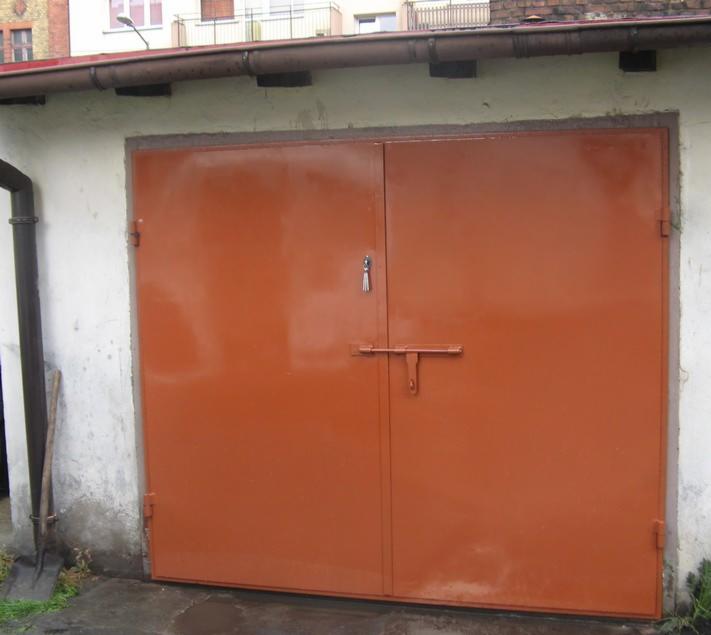 Pomarańczowa, dwuskrzydłowa brama garażowa z prostym zamkiem i zasuwą, zamontowana w garażu z otynkowanymi ścianami. Widoczny fragment rynny i szpadel oparty o ścianę.