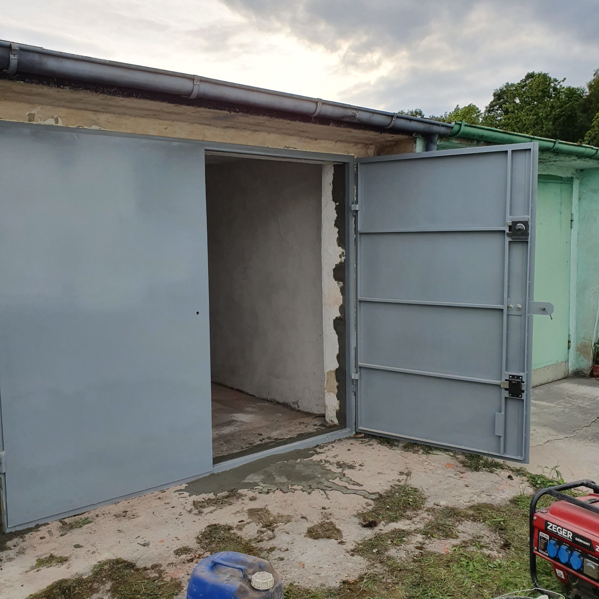 Otwarta szara brama garażowa z widocznym surowym wnętrzem, świeżo osadzona w betonowym podłożu, z generatorem i kanistrem w tle.